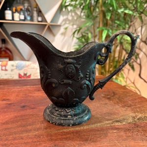 May include: A black metal creamer pitcher featuring an ornate embossed pattern with scrollwork and a central shield-like emblem. The pitcher has a curved handle, a wide pouring spout, and a circular pedestal base with visible signs of wear.