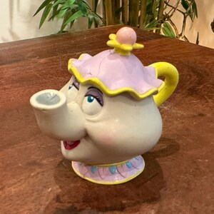 Könnte beinhalten: Eine Keramik-Teekanne in Form einer Figur mit Gesicht, bestehend aus einem weißen Körper, einem gelben Henkel und einem lavendelfarbenen Deckel mit gelber Blumenverzierung. Die Kanne hat aufgemalte blaue Augen, violette Lider und einen roten Mund.