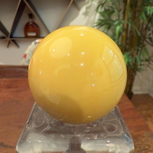 May include: A large, spherical, yellow glass object, possibly a decorative element or paperweight, is displayed on a clear, geometric glass base. The sphere reflects light, creating highlights. The background includes a wooden surface and a glimpse of a shelf.