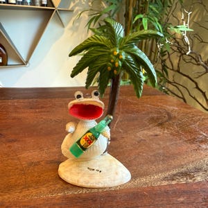 May include: A whimsical frog figurine with a wide red mouth, holding a green bottle, and standing on a seashell. A green palm tree is behind the frog. The figurine is a decorative item.