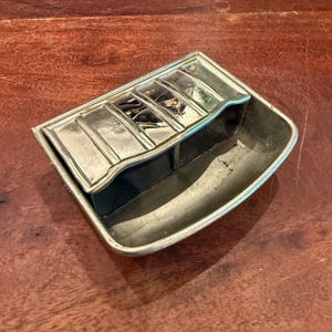 May include: A vintage, rectangular ashtray with a silver-colored metal finish. The ashtray features a divided design with a raised section for resting items and a lower compartment. The piece is set on a wooden surface.