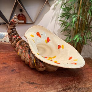 May include: A ceramic decorative shell-shaped bowl with a brown, cream, and orange color scheme. The bowl has a textured, spiraling tail and is decorated with colorful speckles. The bowl is placed on a wooden surface.