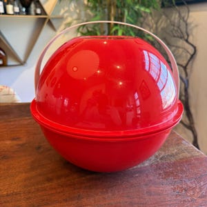 Puede incluir: Una cubitera esférica roja brillante con un asa transparente. La cubitera está hecha de un material brillante y tiene un diseño de dos partes. El asa se arquea sobre la parte superior y la cubitera se encuentra sobre una superficie de madera.