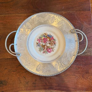 May include: A silver-toned metal serving tray with two handles. The tray has a hammered texture and a decorative floral pattern around the rim. A smaller, round plate with a floral and fruit design sits in the center.