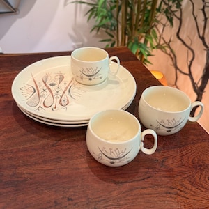 May include: A set of cream-colored ceramic dinnerware featuring three round plates stacked together and two matching mugs. The pieces are decorated with a brown and gray abstract line art pattern consisting of stylized floral and geometric shapes.