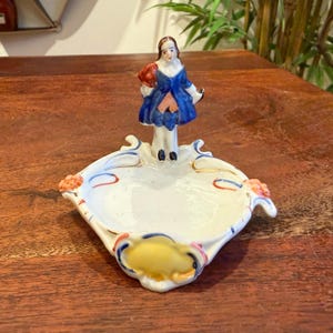 May include: A vintage porcelain trinket dish with a small figurine of a person in a blue dress. The dish is white with gold trim and floral accents. The figurine is holding a red object.