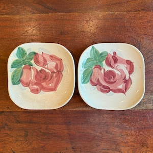 May include: Two square ceramic plates with rounded corners. Each plate features a hand-painted design of a pink rose with green leaves on a cream-colored background. The plates are displayed on a dark wood surface.