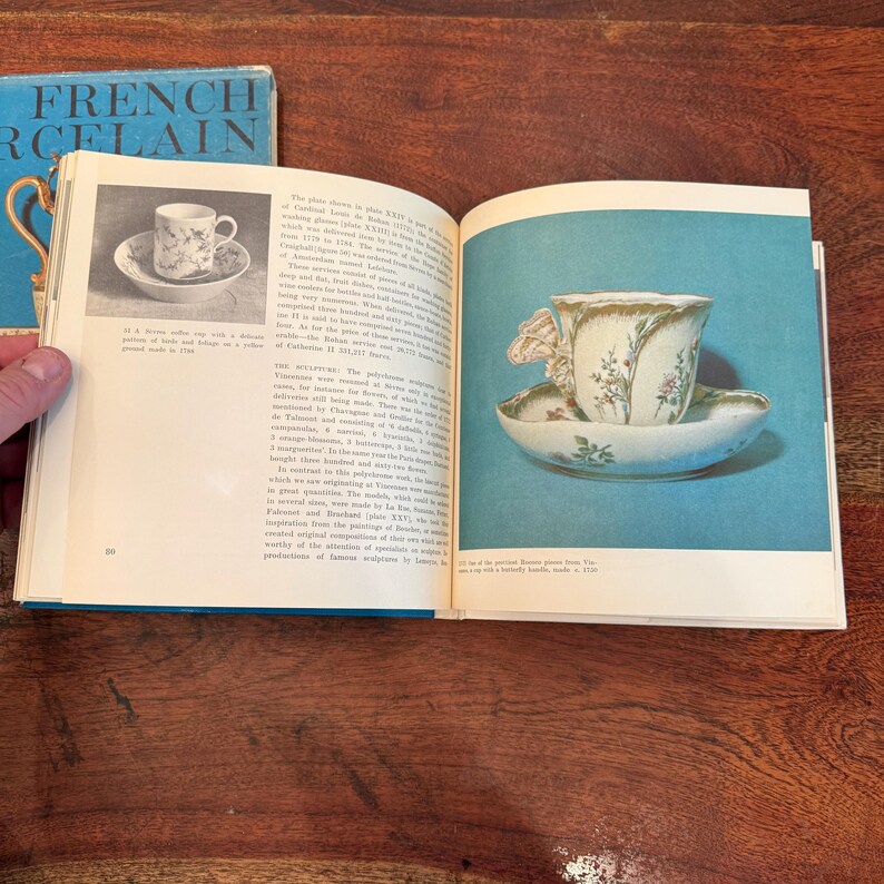 May include: An open book with a blue cover titled "FRENCH PORCELAIN" and a page featuring a vintage teacup and saucer. The teacup and saucer have floral designs and gold trim. The book is open on a wooden surface.