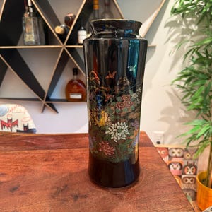 May include: A tall, cylindrical black ceramic vase featuring a detailed, multi-colored floral design. The artwork depicts various chrysanthemums in shades of white, red, yellow, and green, accented with small birds and delicate foliage across the surface.