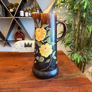 May include: A brown ceramic pitcher with a floral design. The pitcher has a brown glaze with yellow flowers and green leaves painted on it. The pitcher has a wide base and a narrow neck. The pitcher has a handle on the side. The pitcher is signed "Hong" on the bottom.