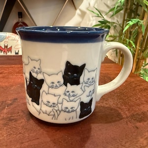 May include: A white ceramic mug featuring a repeating pattern of black and white cat illustrations. The mug has a dark blue rim and a matching dark blue interior. The handle is white and curved, extending from the side of the cylindrical vessel.