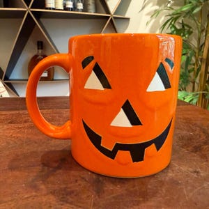 May include: A bright orange ceramic mug shaped like a jack-o'-lantern. The mug features a molded pumpkin face with two triangular eyes and a wide, jagged mouth painted in black and white. It has a curved handle on the left side and a smooth finish.