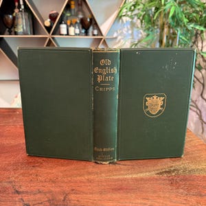 Peut inclure: Un livre ancien intitulé "Old English Plate" par Cripps, troisième édition. Le livre est relié en vert foncé avec des lettres dorées et un emblème doré sur la couverture. Le livre est ouvert, révélant le dos et la couverture intérieure.