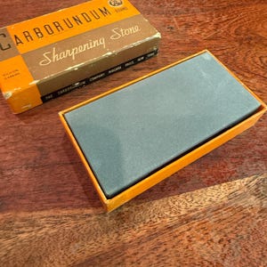 May include: A vintage Carborundum brand sharpening stone in its original box. The box is orange and brown with the words "Carborundum Sharpening Stone" printed on it. The sharpening stone is gray and rectangular, resting in an orange box.