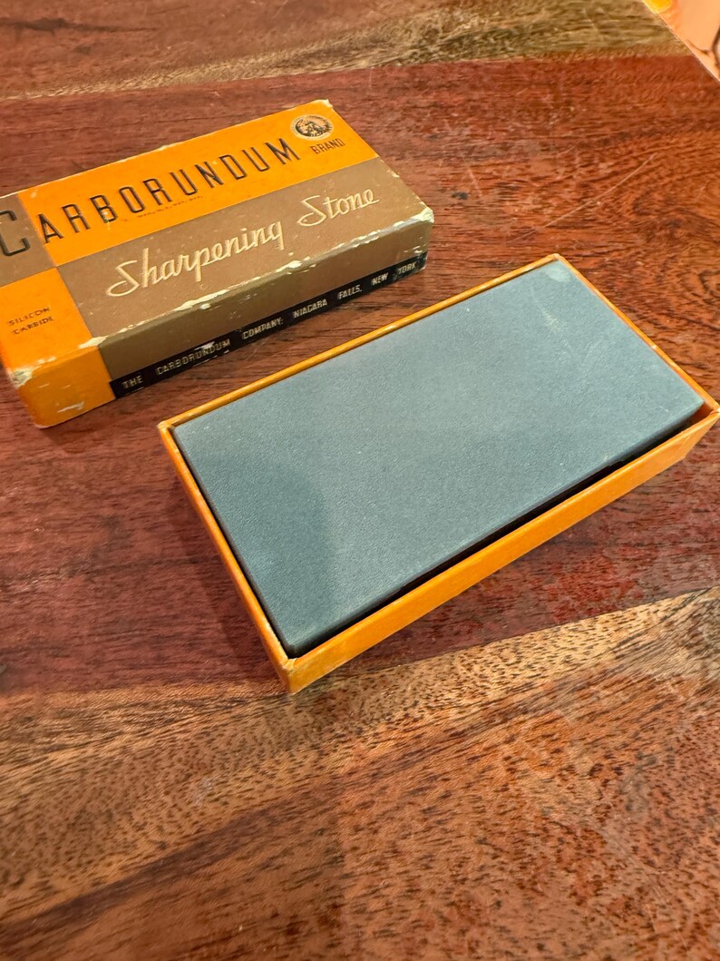 May include: A vintage Carborundum brand sharpening stone in its original box. The box is orange and brown with the words "Carborundum Sharpening Stone" printed on it. The sharpening stone is gray and rectangular, resting in an orange box.