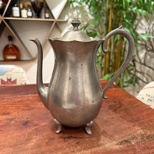 May include: A vintage silver-colored metal coffee pot with a lid, curved handle, and three small feet. The pot has a long, curved spout and a decorative finial on the lid. The pot is sitting on a wooden surface.