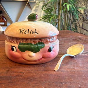 May include: A vintage ceramic relish jar with a smiling face design, featuring a tan lid with the word "Relish" and a green olive-shaped knob. A silver spoon rests beside the jar. The jar has pink cheeks and a green leaf-shaped accent.