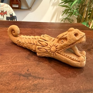 May include: A tan terracotta sculpture of a serpent with an open mouth, visible fangs, and a coiled tail. The body features textured scale patterns and a sun-like circular motif on the side of the head, crafted from natural clay material.