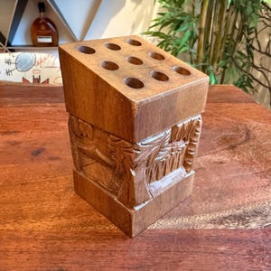 May include: A wooden desk organizer with a rectangular shape and a series of nine circular holes on the top surface. The organizer is made of a light brown wood and features carved relief details on the sides.
