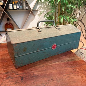 Peut inclure: Une boîte à outils en métal bleu sarcelle vintage avec une finition patinée et une poignée centrale en métal. L'avant présente un emblème circulaire rouge et blanc avec le texte Wards Master Quality. La boîte rectangulaire montre de l'usure.