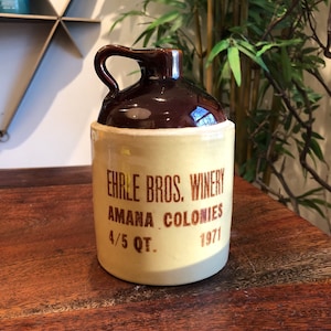 May include: A brown and tan ceramic jug with a handle. The jug has the text "Ehrle Bros. Winery Amana Colonies 4/5 Qt. 1971" printed on it.