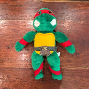 Può includere: Un peluche raffigurante una tartaruga verde con una maschera rossa sugli occhi, fasce rosse sulle braccia e sulle gambe. La tartaruga ha un piastrone giallo e una cintura grigio scuro con una fibbia marrone con la lettera R ricamata in giallo.
