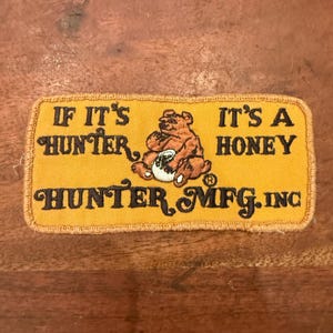 Puede incluir: Un parche rectangular bordado en amarillo que muestra un oso pardo sosteniendo una olla blanca. El texto dice IF IT'S HUNTER, IT'S A HONEY y HUNTER MFG. INC en letras negras. El parche tiene un borde cosido y una superficie de tela texturizada.
