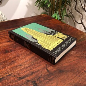 May include: A black hardback book titled "Lands and Peoples" with a green and yellow illustration of a city skyline on the cover. The book is lying on a wooden surface.