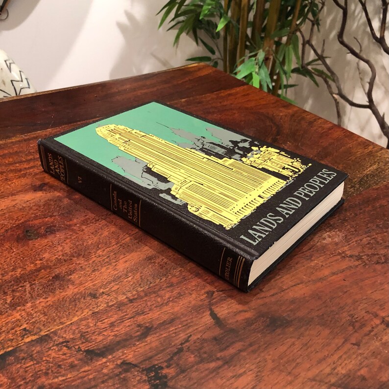 May include: A black hardback book titled "Lands and Peoples" with a green and yellow illustration of a city skyline on the cover. The book is lying on a wooden surface.