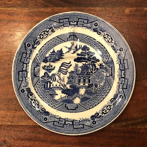 May include: A round, blue and white ceramic plate with a Willow pattern design. The plate features a detailed landscape scene with pagodas, trees, and a bridge. The rim has a decorative border.