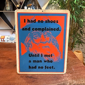 Peut inclure: Panneau rectangulaire avec un fond bleu et une bordure orange. Le panneau présente un portrait d'un homme barbu et le texte "I had no shoes and complained. Until I met a man who had no feet."