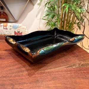 May include: A rectangular ceramic tray with a glossy black finish and wavy, raised edges. The rim features iridescent metallic highlights in shades of bronze, green, and blue, creating a reflective surface that catches the light along the curved borders.