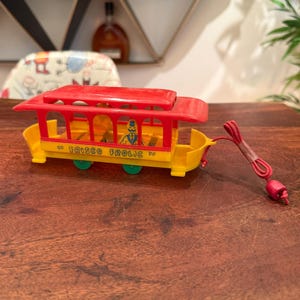 May include: A vintage plastic toy trolley with a bright red roof and yellow body. The side features the text FRISCO FROLIC in black lettering. It includes a small yellow figure inside, green wheels, and a red pull string with a plastic bead end.