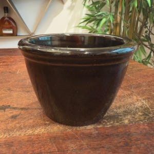 May include: A dark brown ceramic planter with a wide rim and a slightly tapered body. The planter has a glossy finish and sits on a wooden surface. Ideal for indoor or outdoor plants.