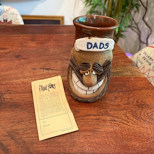 Könnte beinhalten: Keramikbecher mit einem lächelnden Gesicht, in Braun- und Beigetönen. Der Becher hat ein weißes Band mit dem Wort "DADS" in Blau. Ein Etikett ist sichtbar, auf dem die Worte "Mark Hines" aufgedruckt sind.