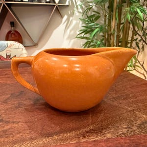 May include: A solid orange ceramic creamer pitcher with a smooth, glossy finish. The pitcher features a rounded body, a curved handle on the left side, and a pointed pouring spout on the right side, creating a simple and functional design.