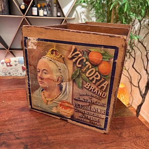 Könnte beinhalten: Eine alte Holzkiste mit einem farbigen Etikett, das ein Porträt von Königin Victoria mit Krone, zwei Orangen an einem Zweig und den Text Victoria Brand, Grown and Packed on Arlington Heights by Victoria Avenue Citrus Association zeigt.