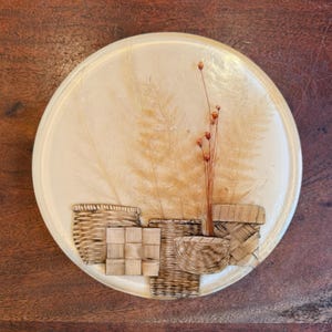 May include: A round, cream-colored decorative tray with a raised border. The tray features a design of woven baskets and dried plants in shades of brown and beige. The tray is set on a dark wood surface.