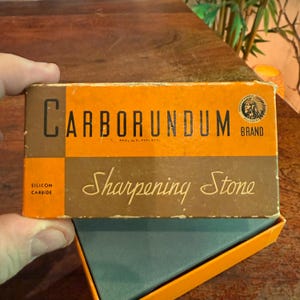 May include: A vintage Carborundum brand sharpening stone in its original box. The box is orange and brown with the words "Carborundum" and "Sharpening Stone" in a classic font. The box also features the brand logo and the words "Silicon Carbide".