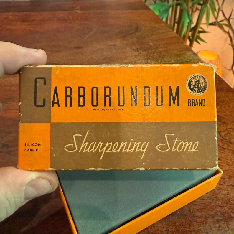 May include: A vintage Carborundum brand sharpening stone in its original box. The box is orange and brown with the words "Carborundum" and "Sharpening Stone" in a classic font. The box also features the brand logo and the words "Silicon Carbide".