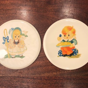 Könnte beinhalten: Zwei antike Keramikteller mit weißem Hintergrund und bunten Illustrationen von Kindern. Ein Teller zeigt ein Mädchen in einem blau-weißen Kleid, das einen Hirtenstab hält, während der andere Teller einen Jungen in einem blau-orangenen Outfit zeigt, der einen Teller mit Essen hält.