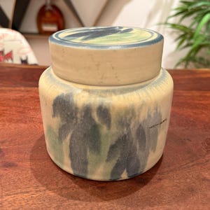 May include: A ceramic jar with a lid. The jar is cylindrical with a cream-colored base and a lid. The jar is decorated with blue and green brushstrokes. The lid has a blue rim and a green and blue design.