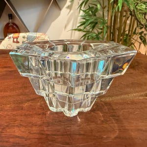 May include: A clear, geometric glass candle holder with a square top and a tiered, faceted design. The candle holder is made of thick glass and sits on a wooden surface. The overall shape is a modern, decorative piece.
