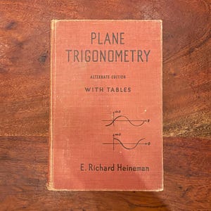 May include: A hardcover textbook titled Plane Trigonometry Alternate Edition With Tables by E. Richard Heineman. The cover is a faded orange color with black text and two mathematical sine and cosine wave graphs printed on the front.