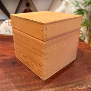 May include: A square wooden box with a removable lid, featuring visible light brown wood grain and interlocking finger joints at each corner. The box has a smooth, flat top surface and straight vertical sides, constructed from natural timber.