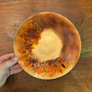May include: A round ceramic plate featuring a smooth, light beige center that transitions into a textured, mottled brown rim. The surface is decorated with a vibrant, swirling pattern of orange, red, and black glaze accents along the left side.