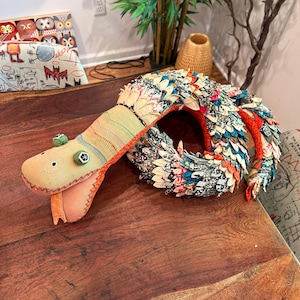 May include: A handcrafted fabric snake toy with a beige head, green eyes, and a colourful patterned body. The snake is coiled on a wooden surface, showcasing a unique, whimsical design. The snake's body is made of various fabric scraps.