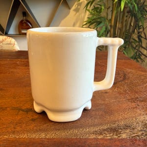 May include: A cream ceramic mug with a distinctive handle and base. The handle has a decorative top, and the base has a scalloped edge. The mug sits on a wooden surface, giving a rustic or vintage feel.