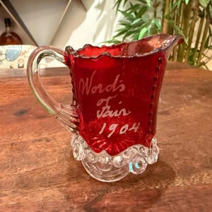 Peut inclure: Un pichet en verre rouge rubis avec une anse en verre transparent et une base en verre transparent à texture perlée. L'avant du pichet est gravé du texte blanc "World's Fair 1904" en écriture cursive, et le bord présente une forme festonnée.