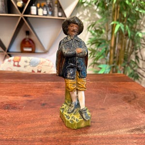 May include: A vintage ceramic figurine of a man in historical attire. The figure wears a dark coat, yellow breeches, and a wide-brimmed hat. The figurine stands on a green base, with arms crossed. The background includes a wooden shelf with bottles.
